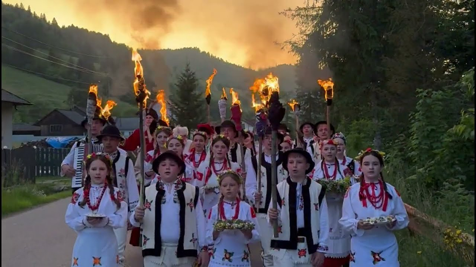 Kinder und Erwachsene in Tracht gehen mit brennenden Fackeln über einen Dorfweg, in der Dämmerung