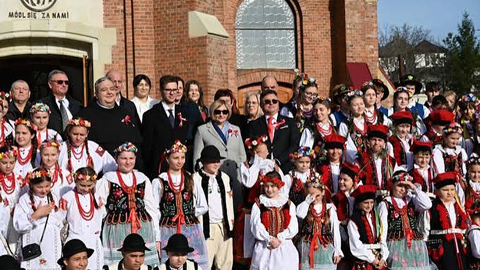 Polska społeczność w strojach ludowych przed kościołem z napisem „Módl się za nami”