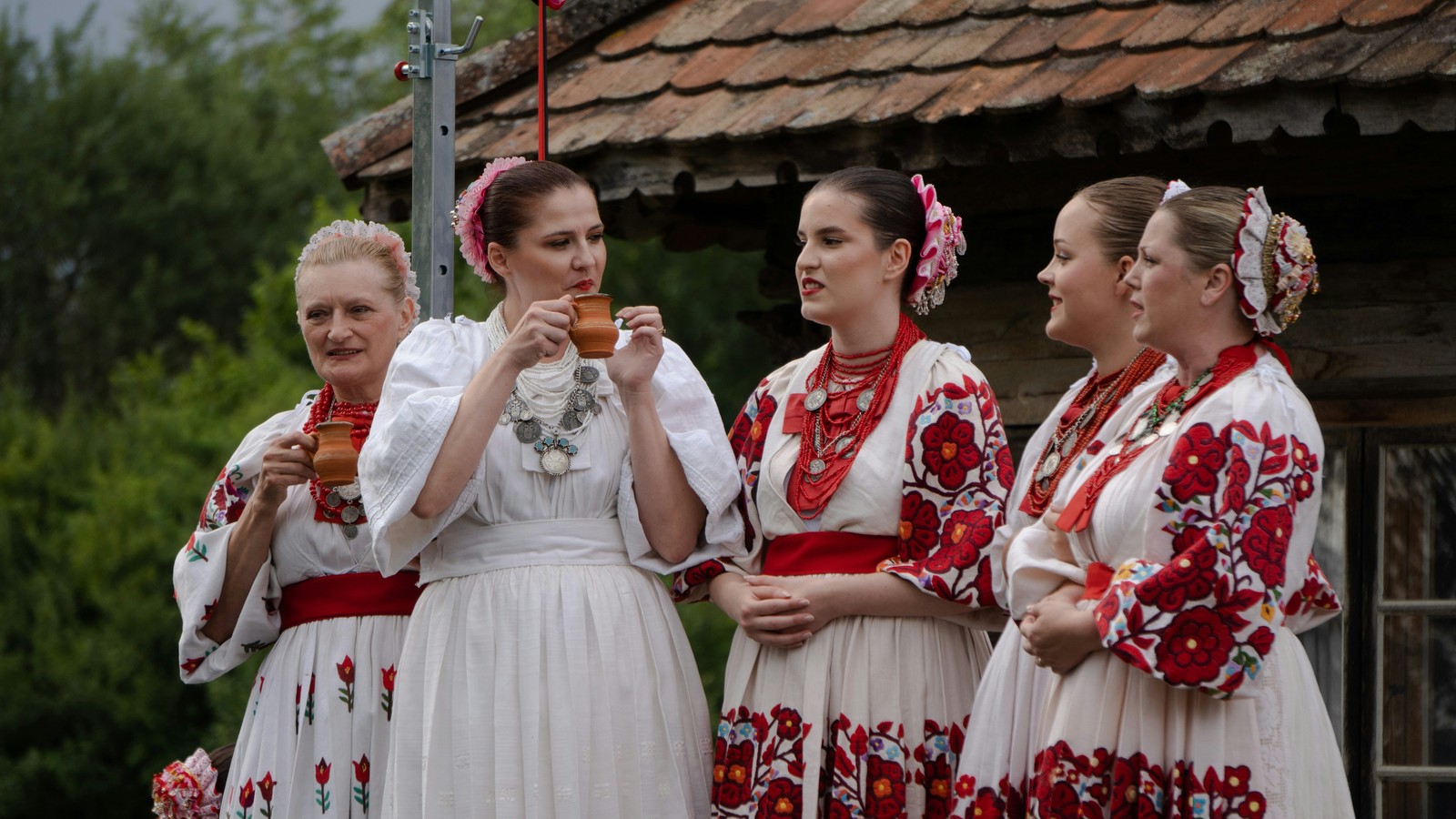 Kobiety w haftowanych strojach ludowych na święcie, przed drewnianą chatą